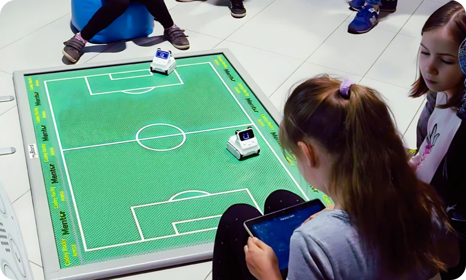 Children controlling Codey Rocky robots on a tabletop soccer pitch.