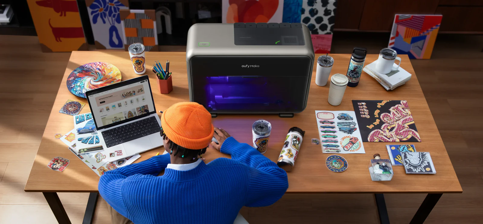 Maker using a laptop beside a eufyMake E1 UV printer, surrounded by printed panels, drinkware and sample designs.