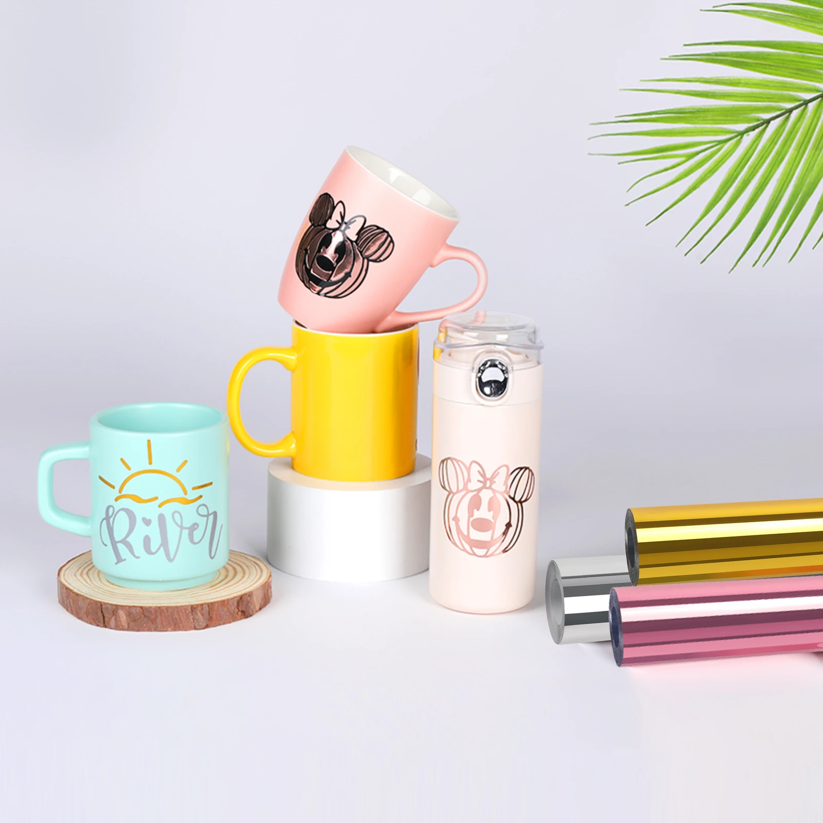 Căni și o sticlă decorate, lângă role de folie tip oglindă aurie, argintie și auriu roz.