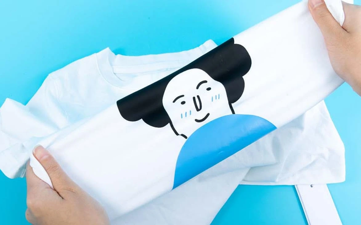 Două mâini întind materialul unui tricou alb cu un desen de față ilustrată din folie termotransfer.