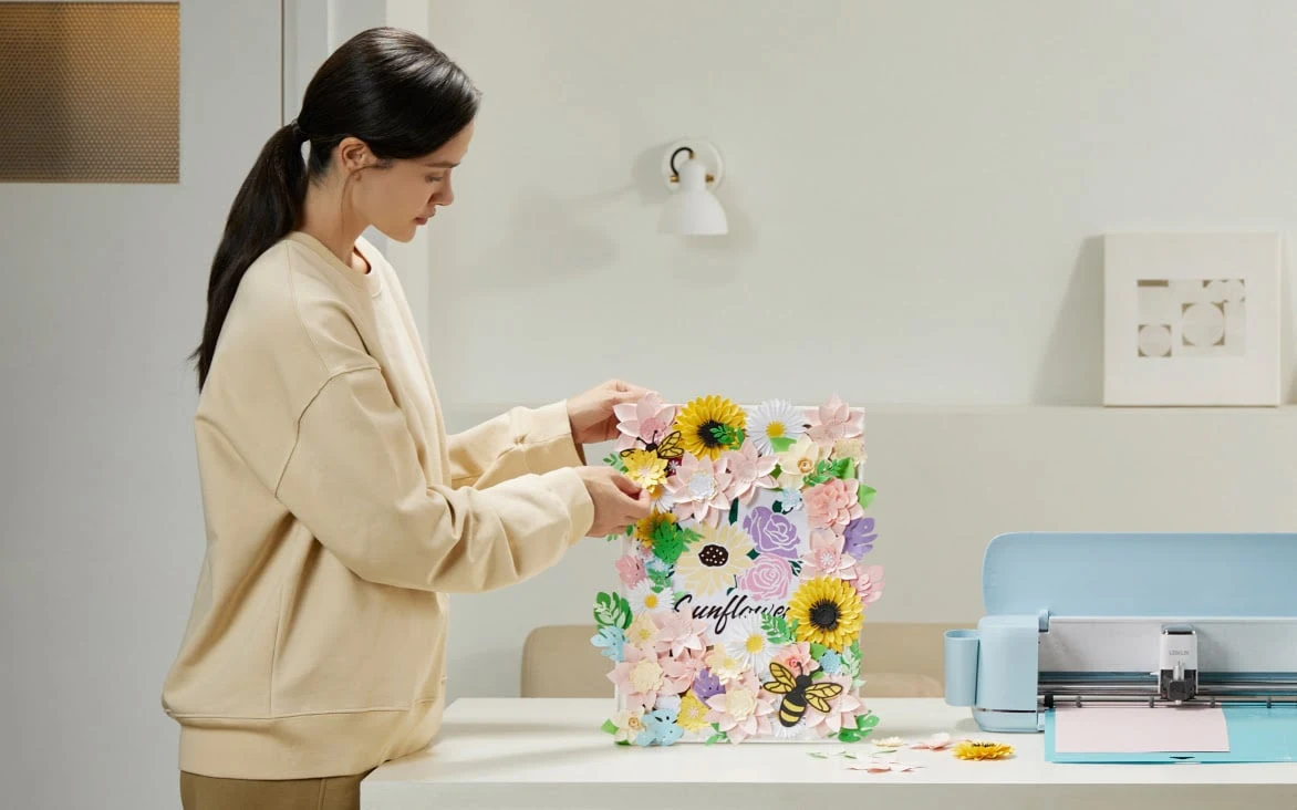 Woman assembling a paper-flower display beside Bubble Blue iCraft on a bright studio desk.