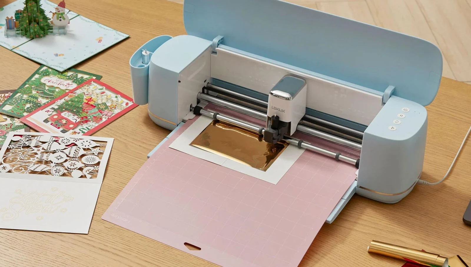 Bubble Blue iCraft working on a gold foil card design on a pink mat, with finished paper projects beside it.