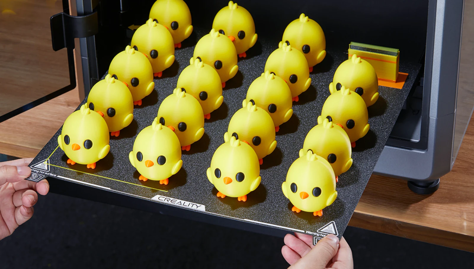 Hands lifting a K2 Plus build plate with rows of multicolour yellow chick prints.