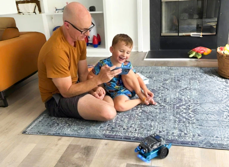 Father and child interacting with mBot2 on a carpet.