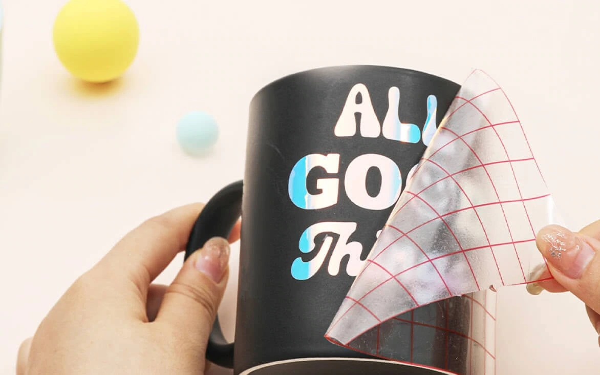 Two female hands holding a black mug with a holographic vinyl design of the text 'All GOOD Things'. A piece of grid transfer tape is being peeled off.