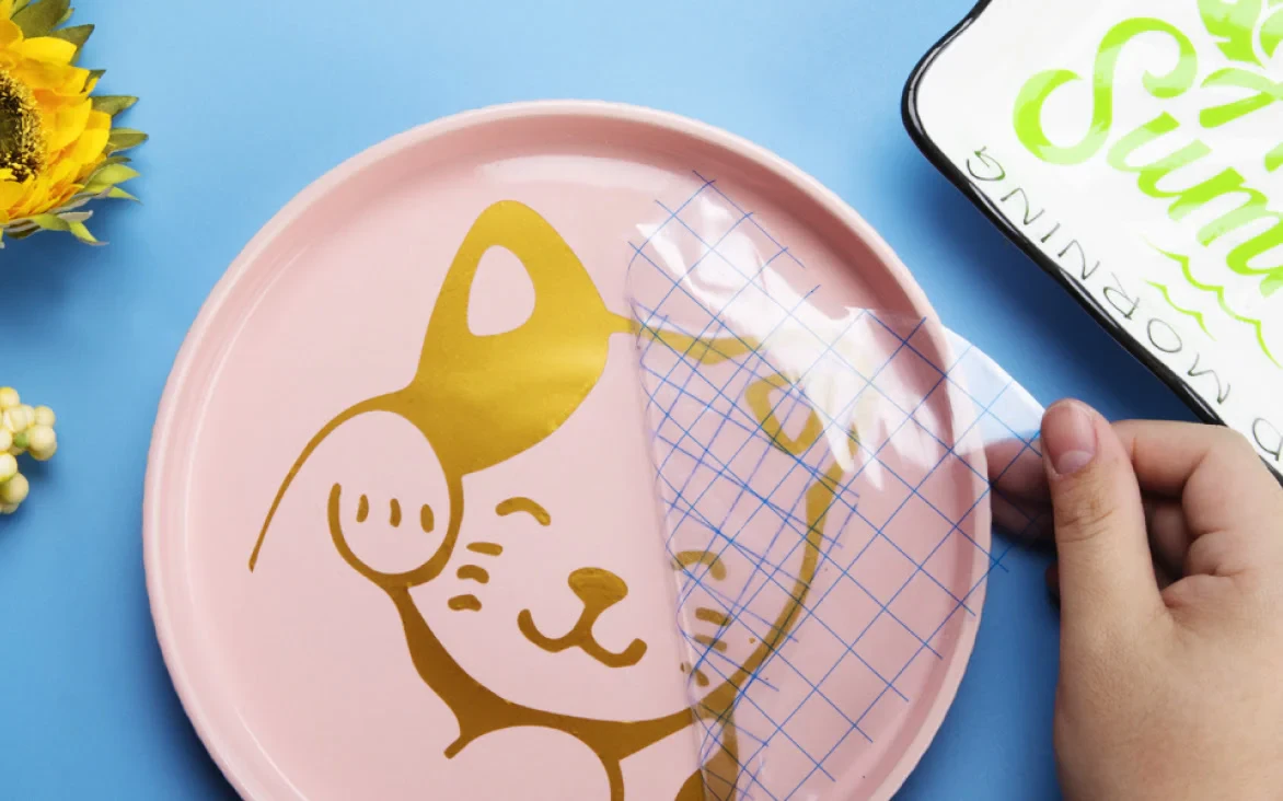 Applying a cat-shaped adhesive vinyl design to a pink plate with transfer tape.